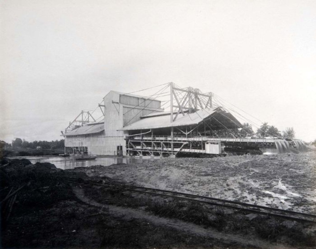 A black-and-white photograph of a large floating tin dredge — a building-sized machine on a barge — at work in shallow water around 1928.
