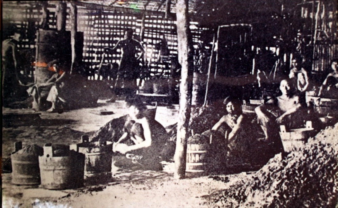 A black-and-white photograph from 1905 of Chinese tin miners working a gravel pit in Phuket, with wooden tools and woven hats.